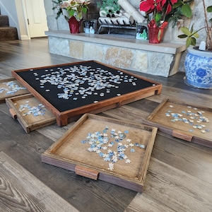 May include: A wooden puzzle table with a black felt surface, surrounded by four wooden trays. The table and trays are filled with puzzle pieces. The table is square, and the trays have wooden handles.