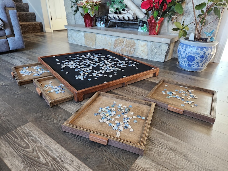 May include: A wooden puzzle table with a black felt surface, surrounded by four wooden trays. The table and trays are filled with puzzle pieces. The table is square, and the trays have wooden handles.
