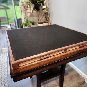 May include: A square, wooden game table with a black playing surface and a brown wooden frame. The table is set on a dark brown table with a lower shelf. The table is approximately 76 cm square.