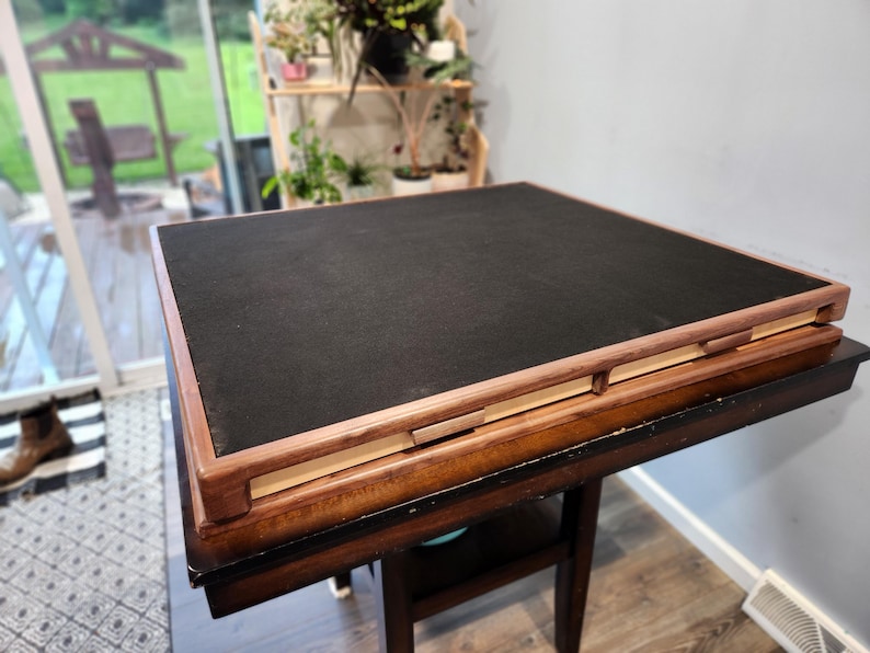 May include: A square, wooden game table with a black playing surface and a brown wooden frame. The table is set on a dark brown table with a lower shelf. The table is approximately 76 cm square.