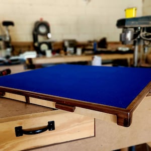 May include: A rectangular wooden frame with a dark blue felt surface. The frame has a light-colored wood border with dark brown accents. The item is on a wooden workbench, suggesting a handcrafted item.