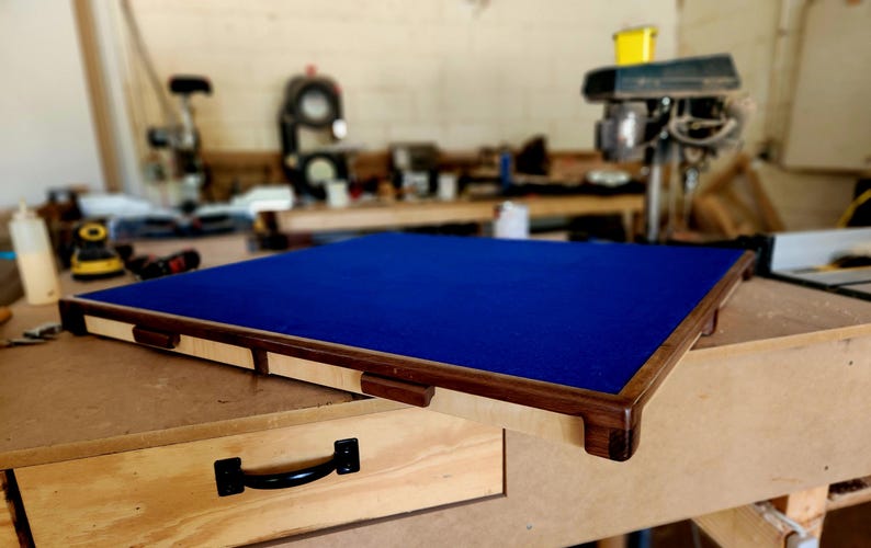 May include: A rectangular wooden frame with a dark blue felt surface. The frame has a light-colored wood border with dark brown accents. The item is on a wooden workbench, suggesting a handcrafted item.