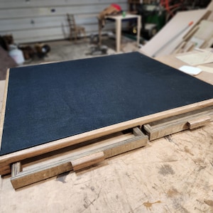May include: A wooden desk with a black felt top and two pull-out drawers. The desk is made of light-colored wood and sits on a wooden surface. The drawers have a simple, rectangular design.