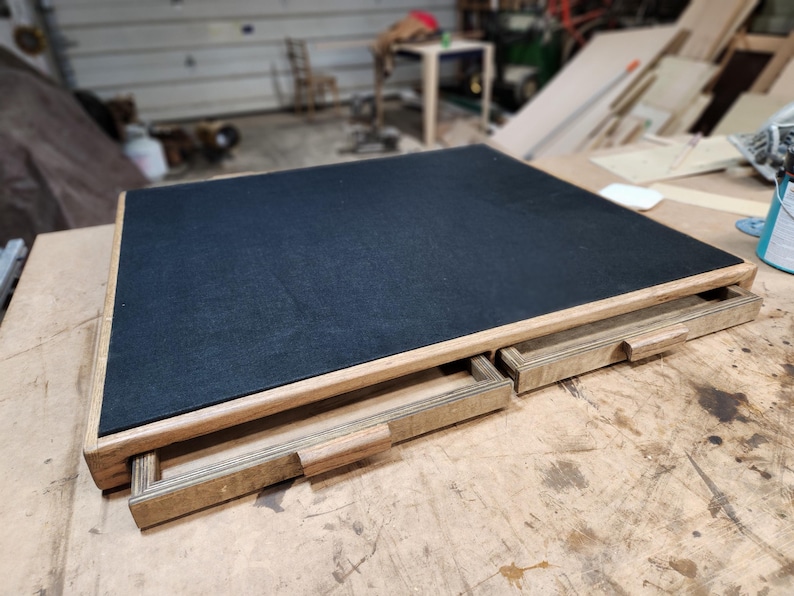 May include: A wooden desk with a black felt top and two pull-out drawers. The desk is made of light-colored wood and sits on a wooden surface. The drawers have a simple, rectangular design.