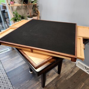 May include: A square wooden game table with a black playing surface and four pull-out drawers. The table has a dark brown frame and sits on a smaller, dark brown table. The drawers are light-coloured wood.