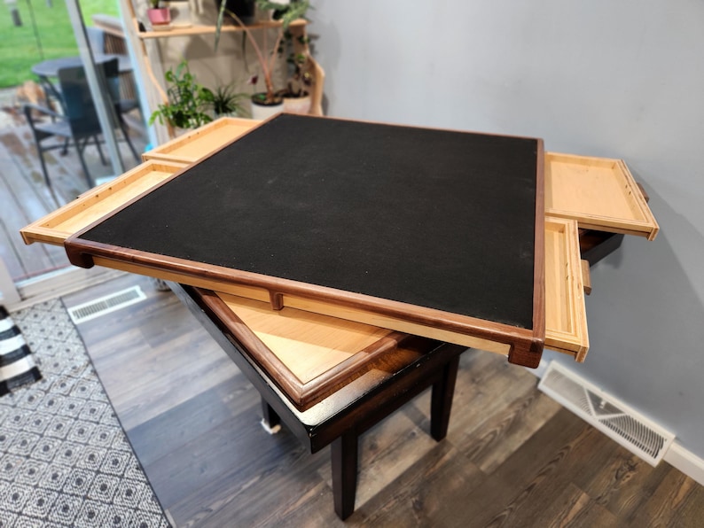 May include: A square wooden game table with a black playing surface and four pull-out drawers. The table has a dark brown frame and sits on a smaller, dark brown table. The drawers are light-coloured wood.