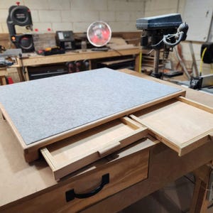 May include: A wooden desk with a gray felt top and two pull-out drawers. The desk is made of light-colored wood and has a simple, functional design. The drawers are open, revealing the interior.
