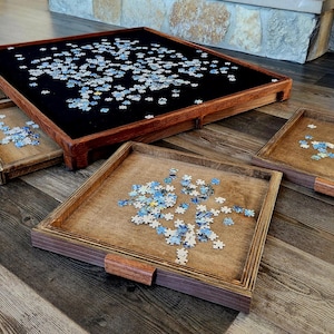 May include: A wooden puzzle table with a black felt surface, surrounded by four smaller wooden trays. The table and trays are filled with puzzle pieces. The trays have wooden handles.