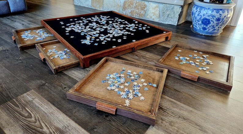 May include: A wooden puzzle table with a black felt surface, surrounded by four smaller wooden trays. The table and trays are filled with puzzle pieces. The trays have wooden handles.