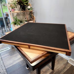 May include: A wooden game table with a black playing surface and a light brown wooden frame. The table is set on top of another table with a lighter wood finish. The table is designed for board games.