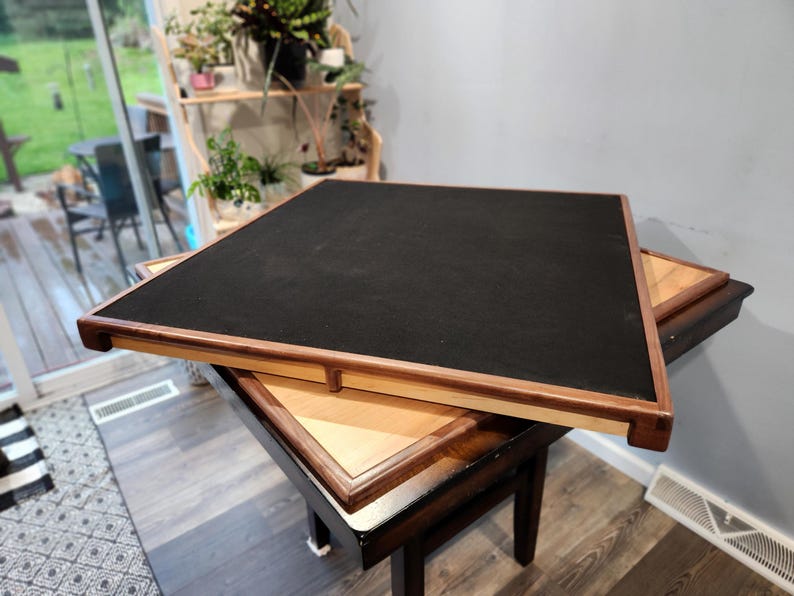 May include: A wooden game table with a black playing surface and a light brown wooden frame. The table is set on top of another table with a lighter wood finish. The table is designed for board games.