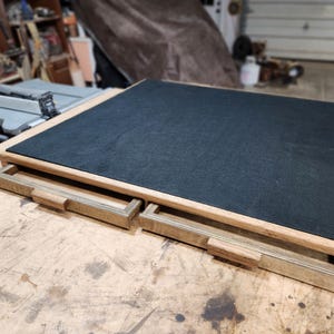 May include: A wooden box with two pull-out drawers. The top surface is covered in black felt. The box has a light brown wooden frame and drawer fronts. The drawers have small, rectangular wooden handles.