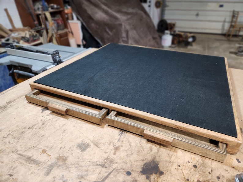 May include: A wooden box with two pull-out drawers. The top surface is covered in black felt. The box has a light brown wooden frame and drawer fronts. The drawers have small, rectangular wooden handles.