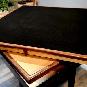 May include: A square wooden game table with a black felt playing surface. The table has a light wood frame with a dark wood border. Another table is partially visible beneath it.