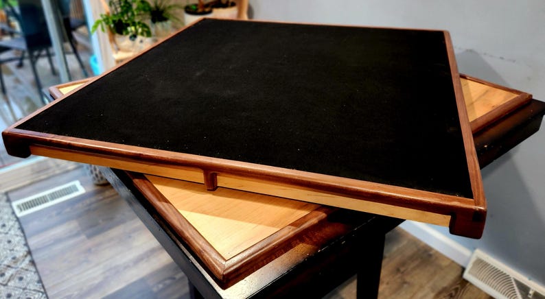 May include: A square wooden game table with a black felt playing surface. The table has a light wood frame with a dark wood border. Another table is partially visible beneath it.