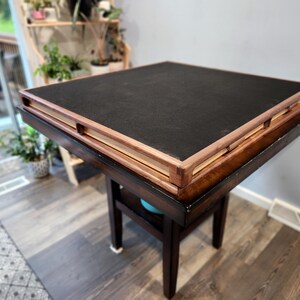 May include: A square, dark brown wooden table with a black top. The table has a lower shelf and four legs. The top has a raised, light brown wooden frame. The table is in a room with a window and plants.