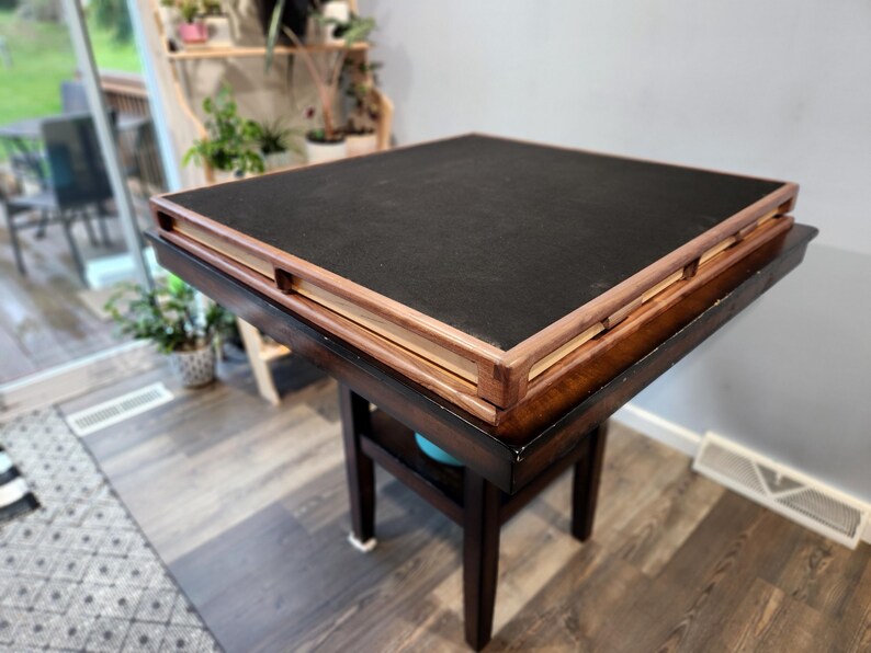 May include: A square, dark brown wooden table with a black top. The table has a lower shelf and four legs. The top has a raised, light brown wooden frame. The table is in a room with a window and plants.