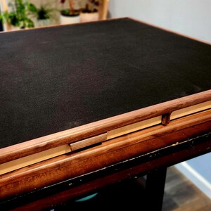 May include: A square wooden game board with a black playing surface. The frame is made of light brown wood, and the board is resting on a dark brown table. The board has a simple, elegant design, with measurements in centimetres.