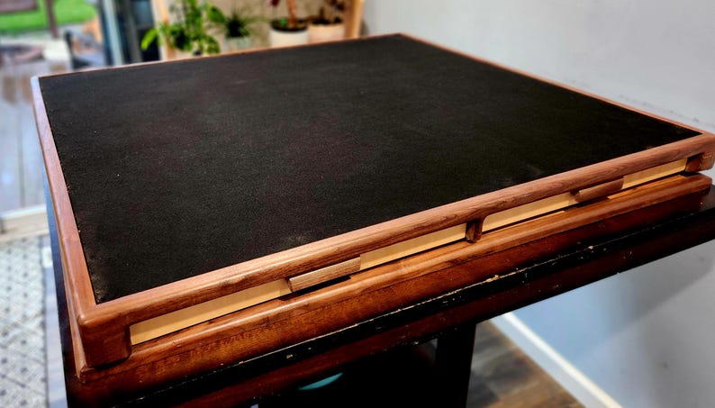 May include: A square wooden game board with a black playing surface. The frame is made of light brown wood, and the board is resting on a dark brown table. The board has a simple, elegant design, with measurements in centimetres.