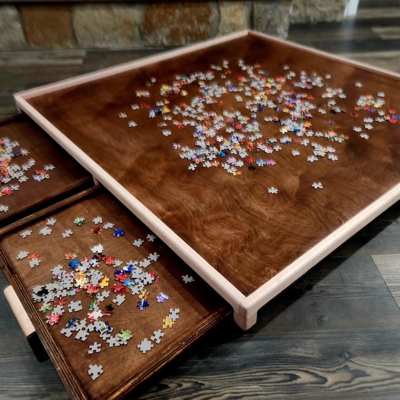 May include: A wooden puzzle table with a dark brown finish and a light-colored frame. The table is partially filled with colorful puzzle pieces, and two pull-out trays are visible, also containing puzzle pieces.