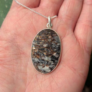 May include: An oval-shaped pendant featuring a textured, multi-colored stone with deep brown, black, and white mineral inclusions. The stone is set in a smooth, polished silver-toned metal bezel and hangs from a delicate, thin silver-toned chain.