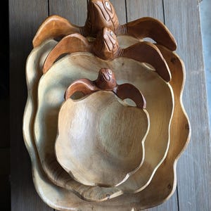 Peut inclure: Ensemble de trois bols gigognes en bois, chacun en forme de fleur avec une figurine de tortue sur le dessus. Les bols sont en bois brun clair avec une finition naturelle. Les tortues sont d'un brun plus foncé.