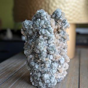以下が含まれることがあります： グレー、白、ピーチ色などの色が混ざった結晶の塊。結晶はざらざらとした質感の表面を持ち、密で不規則な形に配置されています。鉱物標本は木製の表面に展示されています。