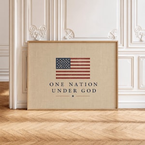 May include: Framed artwork featuring the American flag with red and blue accents. The text "ONE NATION UNDER GOD" is printed below the flag. The artwork is displayed on a light beige background with a wooden frame.