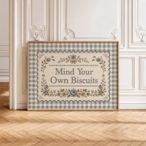 May include: A framed cross-stitch sampler featuring a blue and white gingham background. The center text reads "Mind Your Own Biscuits" in a dark blue font, surrounded by a decorative border of blue and yellow floral motifs and leaves.
