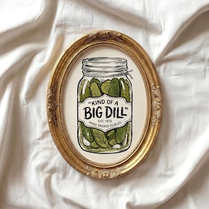 Pode incluir: Uma impressão ovalada com moldura dourada com um frasco de picles. O frasco está rotulado "Kind of a Big Dill" e "Hand-Packed Pickles, Est. 1972". O frasco contém picles verdes, alho e endro. O fundo é um tecido branco texturizado.