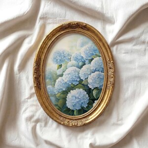 Pode incluir: Pintura ovalada com moldura dourada com flores de hortênsia azuis e folhas verdes. A moldura tem detalhes ornamentados. A obra de arte está sobre um fundo branco. Tema floral.