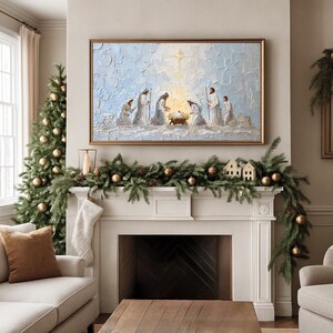 Puede incluir: Una pintura enmarcada que representa la escena de la Natividad cuelga sobre una chimenea decorada con una guirnalda de vegetación, adornos dorados y pequeñas casas. Un árbol de Navidad con adornos dorados está a la izquierda. La pintura tiene un marco dorado.