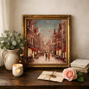 May include: A framed print depicts a romantic European street scene with heart-shaped decorations. The gold frame surrounds the artwork, which features a couple walking down a cobblestone street. A candle, rose, and vase are on a wooden surface.