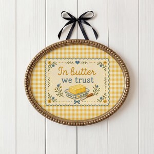 May include: Oval framed cross-stitch art with a yellow gingham background. The artwork features the text "In butter we trust" with a butter dish and butter knife. A black ribbon is tied at the top for hanging.