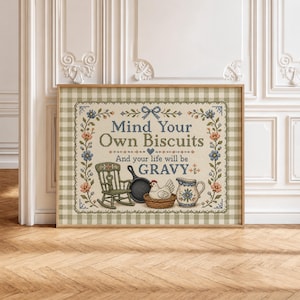 May include: A framed cross-stitch sampler featuring a beige background with a green gingham border. The text reads Mind Your Own Biscuits And your life will be GRAVY. It includes a green rocking chair, a black skillet, a white hen, and a blue pitcher.