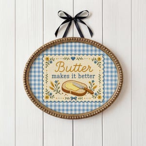 May include: Oval framed wall art with a gingham background and a cross-stitched design. The design features the words "Butter makes it better" with an illustration of buttered toast and a knife. A black ribbon is attached at the top.
