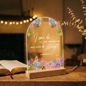 May include: An arched clear acrylic plaque with a wooden base featuring a floral border in shades of blue, purple, and green. The text reads: I can do all this through him who gives me strength Philippians 4:13. The plaque is illuminated.