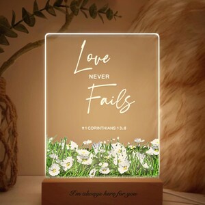 May include: A rectangular LED light with the words "Love Never Fails" and "+1 Corinthians 13:8" in white script. The base features the phrase "I'm always here for you". The bottom of the light has a floral design.