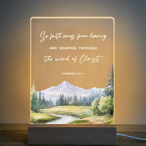 May include: A light-up sign with a watercolour landscape scene of a mountain range, trees, and a river. The sign has the text "So faith comes from hearing and hearing through the word of Christ + Romans 10:17".