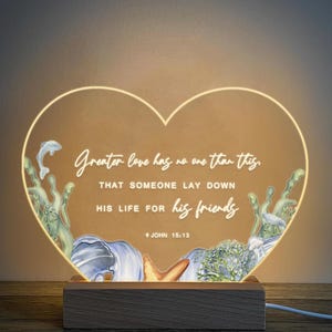 May include: A heart-shaped LED night light featuring the quote "Greater love has no one than this." The design incorporates watercolor illustrations of marine life. The light sits on a wooden base, ideal for home decor.