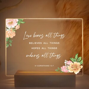 May include: A white acrylic sign with a floral design and the text "Love bears all things, believes all things, hopes all things, endures all things" and the scripture reference "1 Corinthians 13:7".