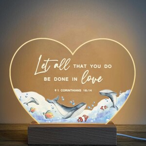 May include: A heart-shaped night light with a wooden base. The light features a watercolour illustration of whales and fish swimming in the ocean. The text "Let all that you do be done in love 1 Corinthians 16:14" is printed on the light.