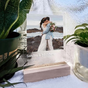 May include: A clear acrylic photo frame with a white wooden base. The frame displays a photo of a couple embracing on a rocky beach. The text "Sandra & Matthew" and the date "02/14/2022" are engraved on the frame.
