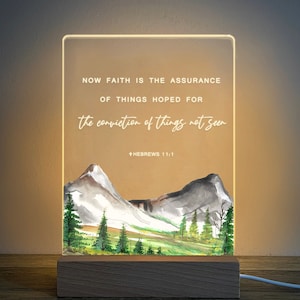 May include: A white acrylic light-up sign with a watercolour mountain scene and the quote "NOW FAITH IS THE ASSURANCE OF THINGS HOPED FOR the conviction of things not seen +HEBREWS 11:1".