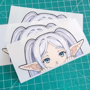 Puede incluir: Tres pegatinas blancas con un personaje de estilo anime con pelo largo y claro en coletas, orejas puntiagudas grandes y ojos azules. El personaje est&aacute; mirando por encima de un borde. Las pegatinas est&aacute;n sobre una esterilla de corte verde.