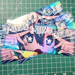 May include: A stack of holographic stickers featuring a stylized anime character with blue hair and star-shaped eyes. The text "MIKU BEAM" is printed across the top of the image. The stickers have a colorful, iridescent finish.