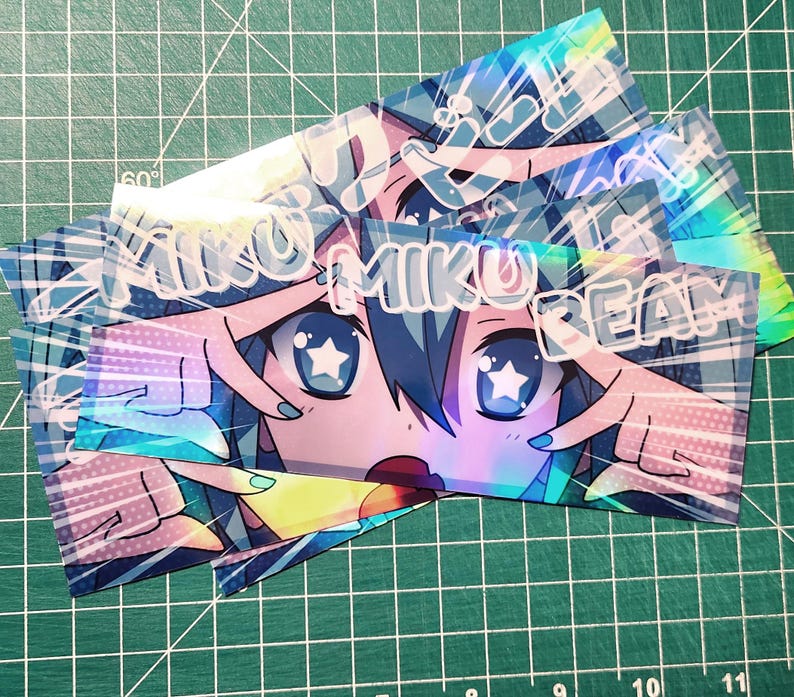 May include: A stack of holographic stickers featuring a stylized anime character with blue hair and star-shaped eyes. The text "MIKU BEAM" is printed across the top of the image. The stickers have a colorful, iridescent finish.