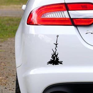 May include: A black and white car decal of a sword in a bonfire. The decal is on the back bumper of a white car.