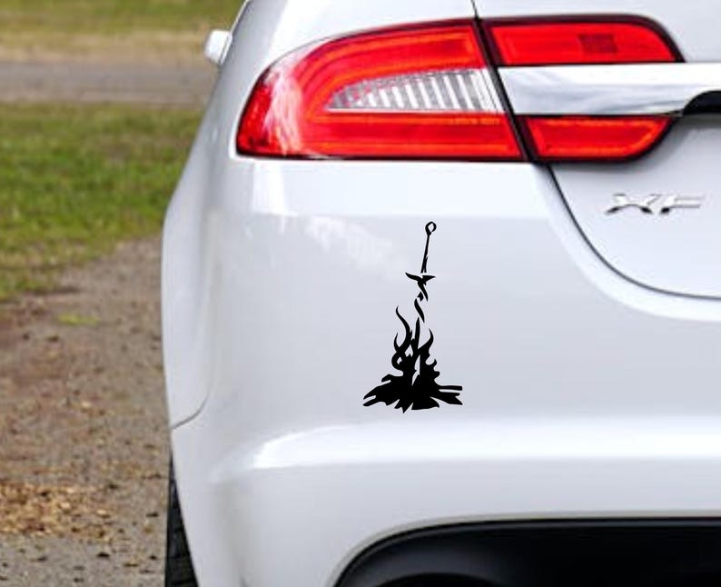 May include: A black and white car decal of a sword in a bonfire. The decal is on the back bumper of a white car.