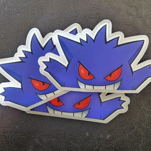 May include: Three layered stickers featuring a purple and blue cartoon ghost with red eyes and a wide grin. The ghost is shown with its arms outstretched.